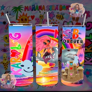 May include: Three colorful, tall, clear plastic cups with straws. The cups have different designs. One cup has a rainbow and a mermaid riding a shark. Another cup has a rainbow and a shark with a sandcastle. The third cup has a rainbow and the text "KAROL G" with a heart.