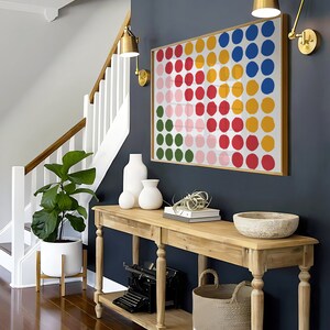 May include: A wooden console table with two shelves, a white ceramic vase, a white ceramic planter with a small plant, a white woven basket, a vintage typewriter, and a bowl on top. The table is against a dark blue wall with a colorful abstract painting of circles.