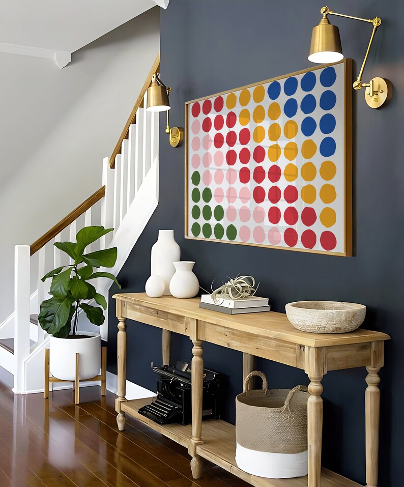 May include: A wooden console table with two shelves, a white ceramic vase, a white ceramic planter with a small plant, a white woven basket, a vintage typewriter, and a bowl on top. The table is against a dark blue wall with a colorful abstract painting of circles.