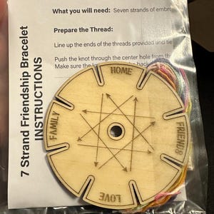 May include: A wooden 7-strand friendship bracelet making kit. The kit includes a wooden disc with slots and the words "HOME", "FAMILY", "FRIENDS", and "LOVE" etched on it. Instructions are included, along with colorful embroidery floss.