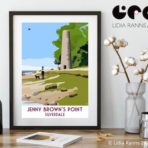 May include: A framed art print showcasing an illustration of Jenny Brown's Point, Silverdale. The artwork includes a person with a dog, a stone tower, and coastal scenery under a blue sky. The print is displayed on a wooden surface.