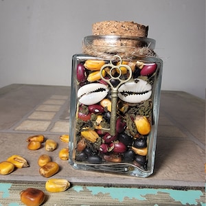 May include: A clear, square glass jar filled with a variety of beans, corn kernels, and dried herbs. A decorative brass key and two white seashells are inside. The jar has a cork stopper and is tied with twine. Some corn kernels are scattered on the surface.