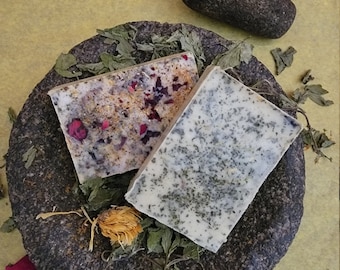 Può includere: Due saponette rettangolari fatte a mano poggiano su un mortaio in pietra grigio scuro, adornate con erbe secche e petali di fiori. Una saponetta è macchiata di petali rossi e scaglie dorate, l'altra con erbe verdi. Un pestello grigio scuro è nelle vicinanze.