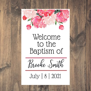 May include: A white sign with pink watercolour flowers at the top. The text reads "Welcome to the Baptism of Brooke Smith July 8 2021".