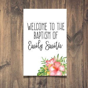 May include: White sign with black text that reads "Welcome to the Baptism of Emily Smith". The sign is decorated with a watercolour floral arrangement of pink and yellow flowers and green leaves.