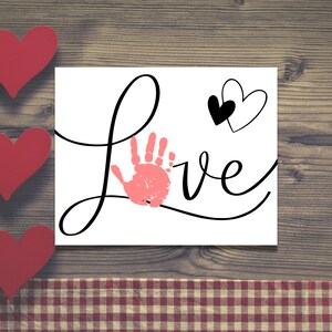 May include: A white card with the word "LOVE" written in black cursive script. A pink handprint is incorporated into the "L" and two black and white hearts are above the word.
