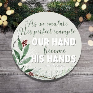 May include: A round, light gray ornament with a white background and green and red floral accents. The ornament features the text "As we emulate His perfect example OUR HAND become HIS HANDS" with the name "DIETER FUCHTDOORF" at the bottom.