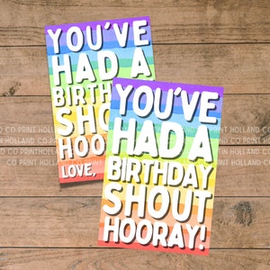 May include: Two rainbow-colored cards with the text "You've had a birthday shout hooray!" in white letters. The cards are on a wood background.