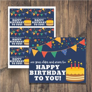 May include: A printable birthday card with a blue background, colorful pennant flags, stars, and a yellow cake with candles. The text reads "one year older and wiser too HAPPY BIRTHDAY TO YOU!"