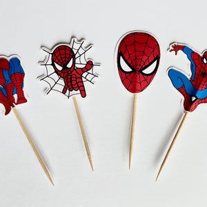 May include: Four Spider-Man themed cupcake toppers. Each topper features a different image of Spider-Man in red and blue, attached to a wooden stick. The background is plain white.