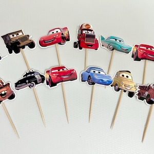 May include: A collection of eleven colorful car-themed cupcake toppers. The toppers feature various cartoon car designs, including a blue tow truck, a red race car, and a yellow vintage car, all attached to wooden skewers.