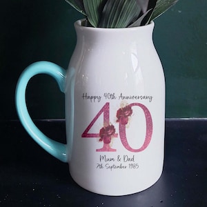 Könnte beinhalten: Eine weiße Keramikvase mit hellblauem Griff, die orangefarbene und gelbe Blumen enthält. Die Vase ist mit dem Text "Happy 40th Anniversary" und "Mum & Dad 7th September 1985" verziert.