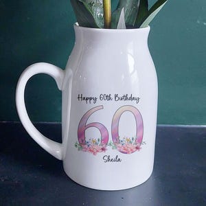 Puede incluir: Un jarrón de cerámica blanco con asa, que contiene vibrantes flores naranjas y amarillas. El jarrón está decorado con el texto "Happy 60th Birthday", el número "60" en rosa y el nombre "Sheila".