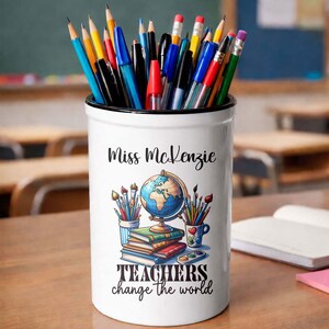 Può includere: Portapenne in ceramica bianca con bordo nero, pieno di matite e penne colorate. Il portapenne presenta il testo "Miss McKenzie" e una grafica di un mappamondo, libri e pennelli. È presente anche la frase "TEACHERS change the world".