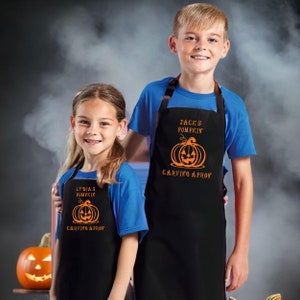May include: Two children wearing black aprons with orange pumpkin designs and the text "Carving Apron". The aprons have brown straps and are worn over blue tops. A carved pumpkin is visible in the background, suggesting a Halloween theme.