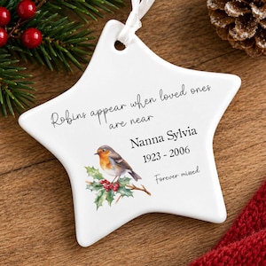 May include: A white star-shaped ceramic ornament featuring a robin illustration on a holly branch. The text reads: Robins appear when loved ones are near, Nanna Sylvia, 1923-2006, Forever missed. It includes a white ribbon for hanging.
