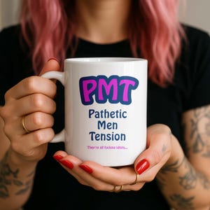 Pode incluir: Caneca de cerâmica branca com o texto "PMT Pathetic Men Tension" em roxo e azul. A caneca também inclui a frase "They're all fucking idiots..." em uma fonte menor.