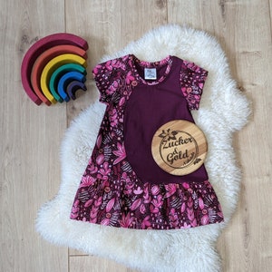 Könnte beinhalten: Ein burgunderfarbenes Kleid mit Blumendruck und einem Rüschen-Saum. Das Kleid hat ein großes, geschwungenes Panel aus einfarbigem Burgunderstoff. Das Kleid liegt auf einem weißen Kunstfell-Hintergrund. Das Kleid hat ein Etikett mit der Aufschrift "Zucker & Gold" darauf.