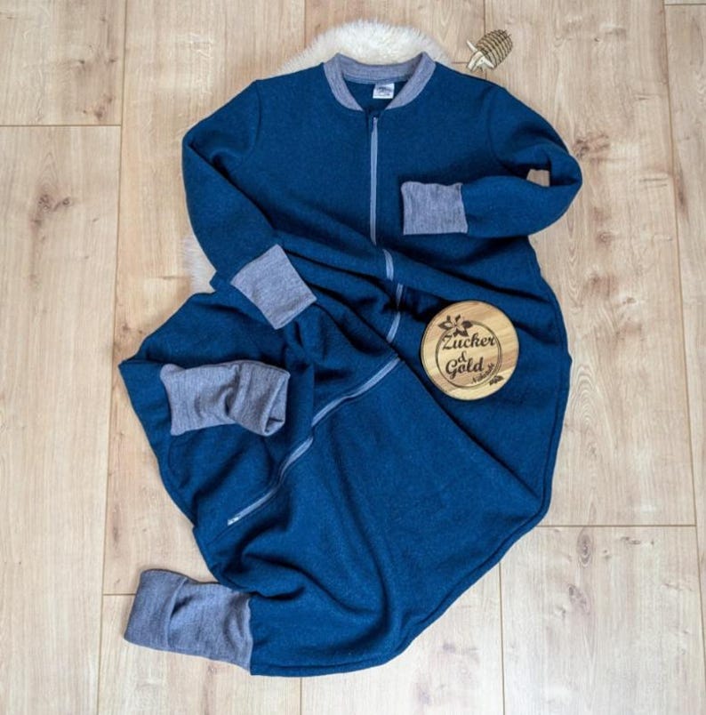 Ein Schlafsack mit Füßen aus weiche Merino-Walk in der Farbe Petrol. Die Bündchen, der Kragen und der Reißverschluss sind grau. Diese Variante hat lange Ärmeln.