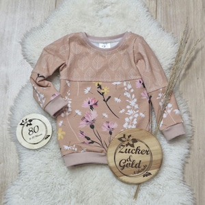 Könnte beinhalten: Ein langärmliger, hellbrauner Pullover mit Blumenmuster. Die obere Hälfte des Pullovers hat ein Muster aus kleinen, weißen Blättern. Die untere Hälfte des Pullovers hat ein Muster aus rosa und weißen Blumen mit grünen Stielen. Der Pullover hat einen runden Ausschnitt und lange Ärmel. Der Pullover liegt auf einer weißen, flauschigen Oberfläche. In der unteren rechten Ecke des Bildes befindet sich ein Holzschild mit dem Text "Zucker & Gold Nähstube". In der unteren linken Ecke des Bildes befindet sich ein rundes Holzschild mit dem Text "80 9-12 Monate".