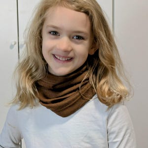 Könnte beinhalten: Ein junges Kind trägt einen braunen Halsschmuck mit einem weißen Langarmshirt.
