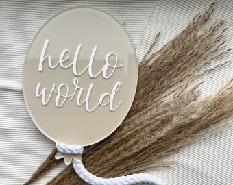 Peut inclure: Une plaque circulaire en acrylique beige avec le texte blanc hello world écrit en lettres cursives. La plaque présente une petite attache blanche en forme de fleur à la base, reliée à un long cordon tressé blanc qui forme une boucle.