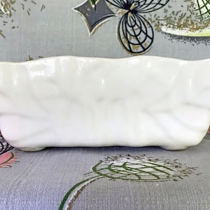 以下が含まれることがあります： 白く長方形のセラミック皿。葉のようなテクスチャパターンがあります。縁はわずかに波打っており、3つの小さな足の上にあります。細長い形状は、サービングまたはディスプレイに適しています。長さ約20cm。