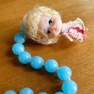 May include: A vintage doll with blonde hair and a red and white dress rests near a strand of light blue beads. The beads are round and strung on a white string, creating a playful and nostalgic aesthetic.