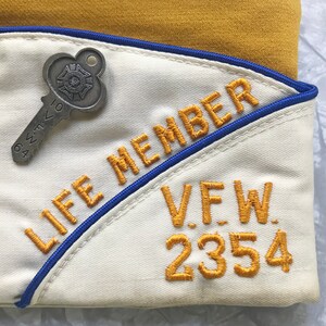Puede incluir: Gorra VFW color crema con texto bordado en oro que dice "LIFE MEMBER" y "V.F.W. 2354". Se adjunta un alfiler plateado en forma de llave. La gorra tiene un ribete azul y est&aacute; sobre un fondo amarillo.
