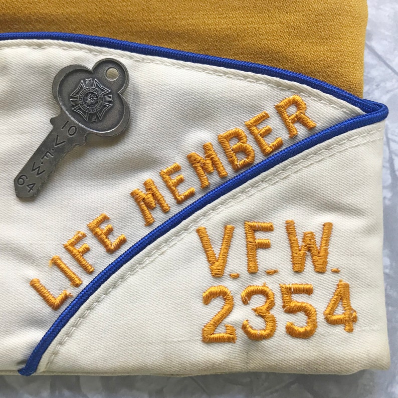 Puede incluir: Gorra VFW color crema con texto bordado en oro que dice "LIFE MEMBER" y "V.F.W. 2354". Se adjunta un alfiler plateado en forma de llave. La gorra tiene un ribete azul y est&aacute; sobre un fondo amarillo.