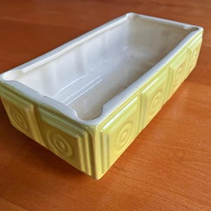 Puede incluir: Jardinera rectangular de cerámica con exterior amarillo pálido e interior crema. Los lados presentan un patrón repetitivo de cuadrados con diseños circulares. La jardinera vacía está sobre una superficie de madera. Objeto decorativo.
