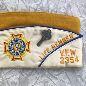 Puede incluir: Un gorro VFW beige y blanco con texto bordado y un alfiler en forma de llave. El gorro presenta las palabras "LIFE MEMBER" y "V.F.W. 2354" en hilo dorado. Tambi&eacute;n hay un emblema de VFW bordado en el gorro.