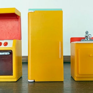 May include: A set of yellow toy kitchen appliances, including an oven with a red interior, a refrigerator with a light blue top, and a sink with a silver faucet. The oven has four red knobs and a dark brown oven door.