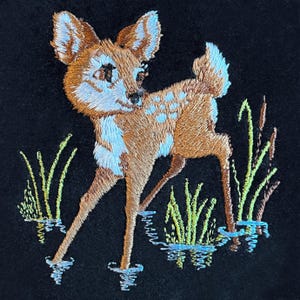 May include: Embroidered image of a fawn standing in water, with a black background. The fawn is brown with white spots and accents. Green and yellow reeds are in the water. The fawn's reflection is visible in the water.