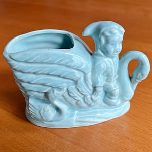Può includere: Un vaso di ceramica azzurra a forma di cigno con una piccola figura seduta sulla schiena. Il vaso ha una grande apertura per le piante ed è appoggiato su una superficie di legno.