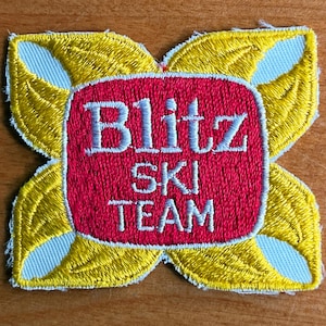 May include: Embroidered patch with a red center and the words "Blitz SKI TEAM" in white. The center is framed by a yellow and light blue floral design. The patch is likely for a ski team.