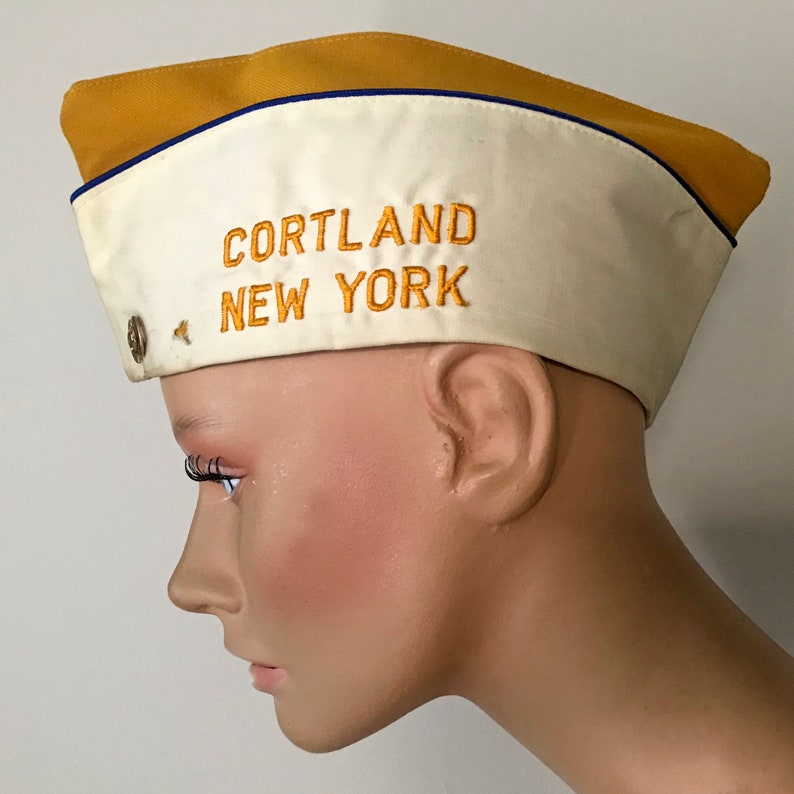 Puede incluir: Gorra de estilo vintage con frente color crema y parte superior amarillo mostaza. La gorra presenta el texto bordado "CORTLAND NEW YORK" en hilo dorado. Un peque&ntilde;o bot&oacute;n de metal est&aacute; en el lateral. La gorra tiene un ribete azul.