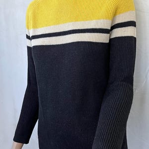 Puede incluir: Un su&eacute;ter de punto acanalado con dise&ntilde;o de bloques de color. La parte superior es amarilla, seguida de una franja blanca, una franja negra y el resto del su&eacute;ter es negro. El su&eacute;ter tiene cuello alto y mangas largas.