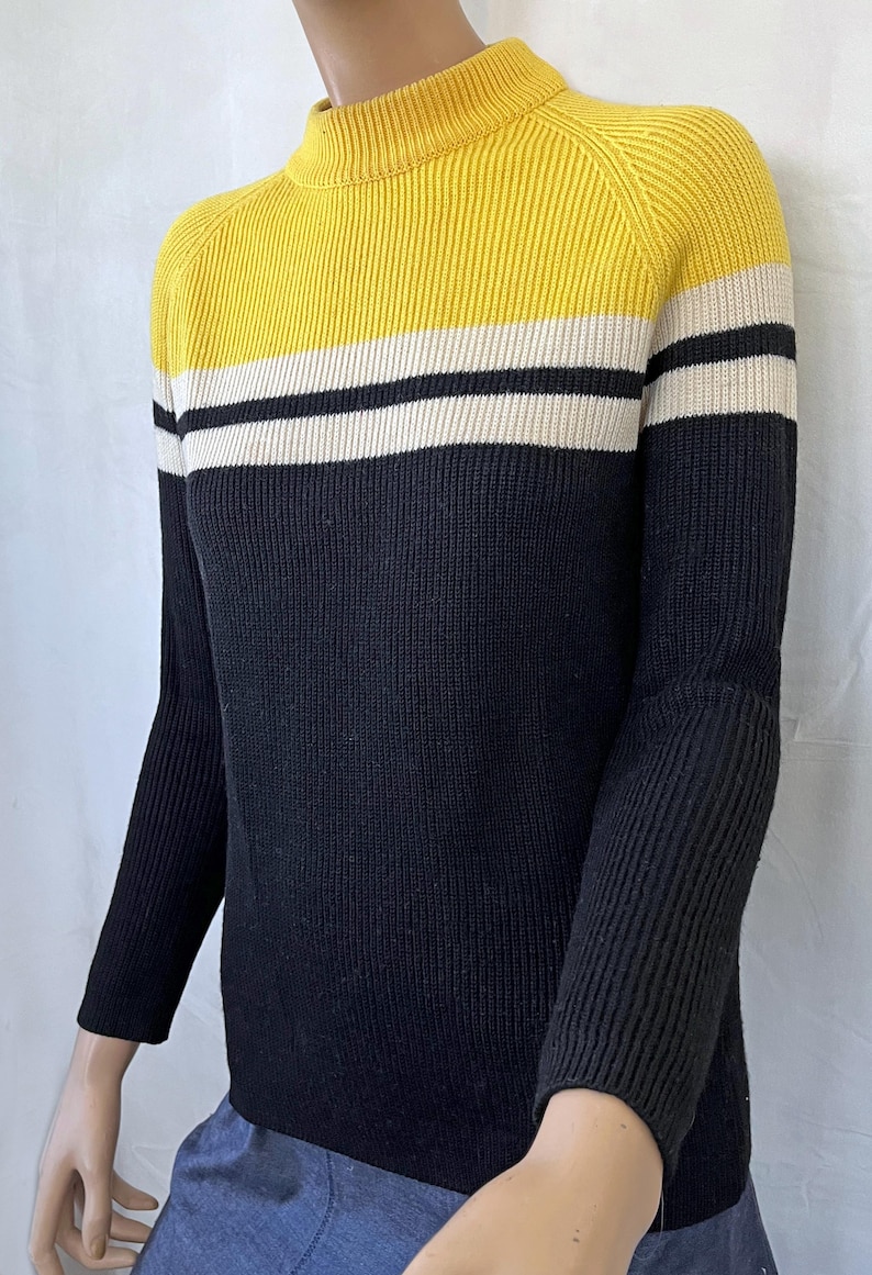 Puede incluir: Un su&eacute;ter de punto acanalado con dise&ntilde;o de bloques de color. La parte superior es amarilla, seguida de una franja blanca, una franja negra y el resto del su&eacute;ter es negro. El su&eacute;ter tiene cuello alto y mangas largas.