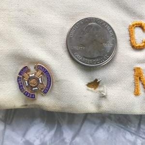 Puede incluir: Primer plano de una tela color crema con un pin de Past Commander dorado y azul, una moneda de veinticinco centavos y letras bordadas en oro. El pin dice "Post 1964-65 2354" y "Past Commander". La moneda es un Quarter Dollar estadounidense.