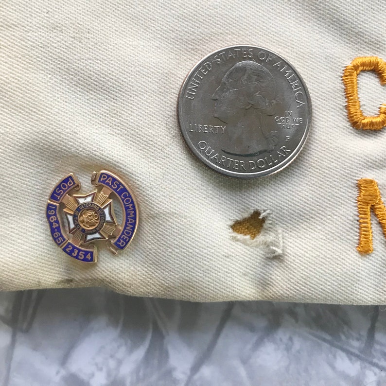 Puede incluir: Primer plano de una tela color crema con un pin de Past Commander dorado y azul, una moneda de veinticinco centavos y letras bordadas en oro. El pin dice "Post 1964-65 2354" y "Past Commander". La moneda es un Quarter Dollar estadounidense.