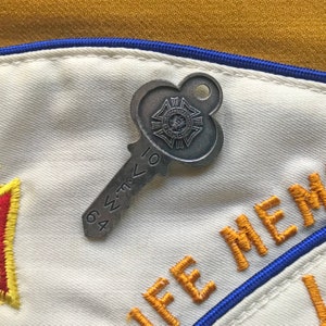 Puede incluir: Un pin plateado en forma de llave con las palabras "Veterans of Foreign Wars" y los n&uacute;meros "64" y "10". El pin est&aacute; sobre una tela blanca con bordados dorados y azules.