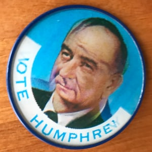 Puede incluir: Un botón de campaña vintage con un retrato de un hombre con traje. El botón tiene un borde azul y un fondo azul claro. Las palabras "VOTE HUMPHREY" están impresas en blanco alrededor del borde.
