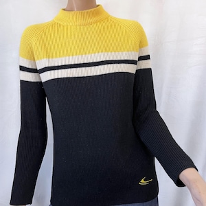 Puede incluir: Un su&eacute;ter de punto acanalado con cuello alto. La secci&oacute;n superior es amarilla, seguida de una franja blanca y negra, y la secci&oacute;n inferior es negra. Un peque&ntilde;o logotipo amarillo est&aacute; en la parte inferior izquierda.