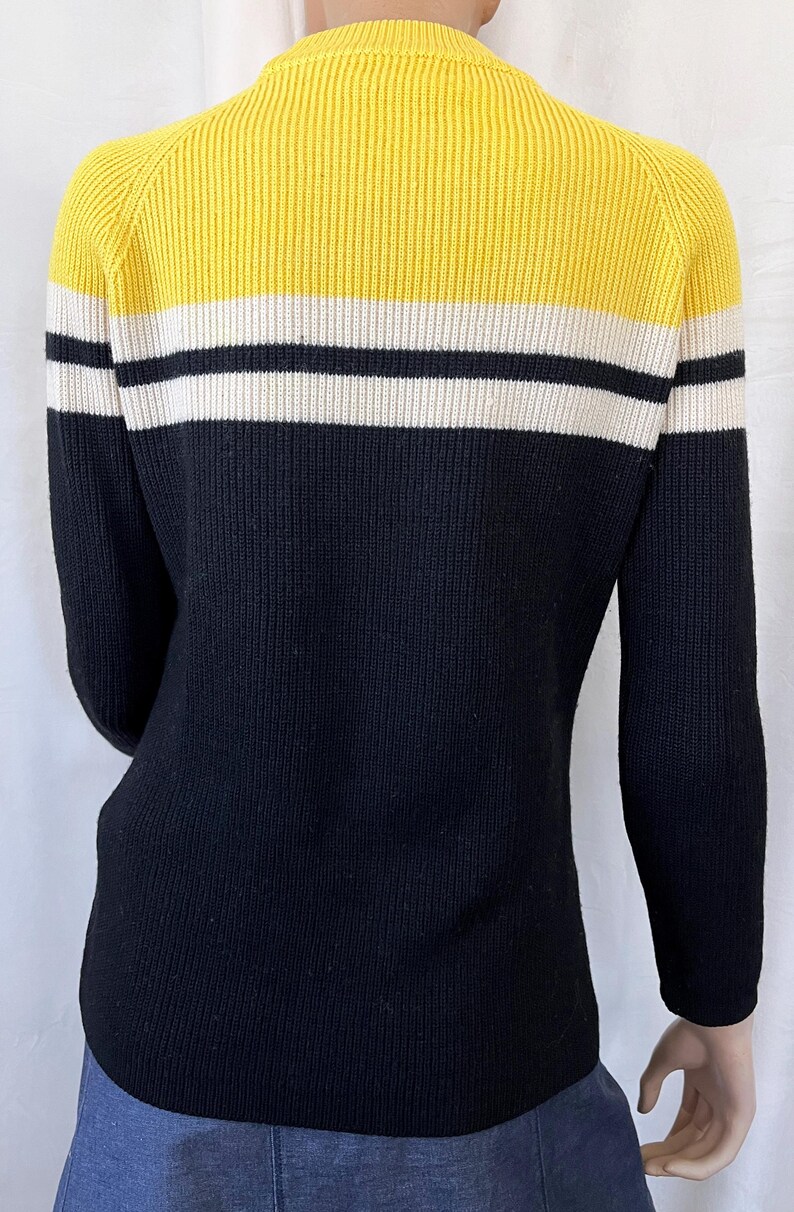 Puede incluir: Un su&eacute;ter de punto acanalado en amarillo, negro y blanco. La parte superior es amarilla, seguida de una franja blanca, una franja negra y el resto del su&eacute;ter es negro. El su&eacute;ter tiene mangas largas.