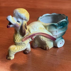 Può includere: Un'antica fioriera a forma di barboncino in ceramica gialla, marrone e verde. Il barboncino tira un carrello con una ciotola per piante. La fioriera ha un design stravagante con una figura di barboncino dettagliata e un carrello decorativo.
