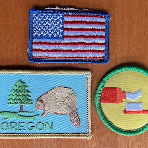 Puede incluir: Tres parches bordados: una bandera americana, un parche rectangular con un castor y la palabra "OREGON", y un parche circular con un cepillo de dientes y pasta dental.