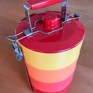 Puede incluir: Una fiambrera vintage de varios niveles en un vibrante degradado de colores. La parte superior y el asa son rojas, pasando por amarillo, naranja y rojo. El asa y los cierres metálicos aseguran los compartimentos apilados. Un recipiente clásico para llevar comidas.