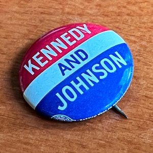 Puede incluir: Un botón de campaña vintage con las palabras "KENNEDY AND JOHNSON" en letras blancas. El botón está dividido en secciones rojas, celeste y azul oscuro. Un alfiler plateado está adherido en la parte posterior.