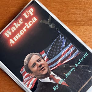Może przedstawiać: Okładka książki z tytułem "Wake Up America" w czerwonym i białym tekście. Okładka przedstawia portret mężczyzny w garniturze i krawacie, z amerykańską flagą w tle. Widoczne jest również nazwisko autora, dr. Jerry'ego Falwella.